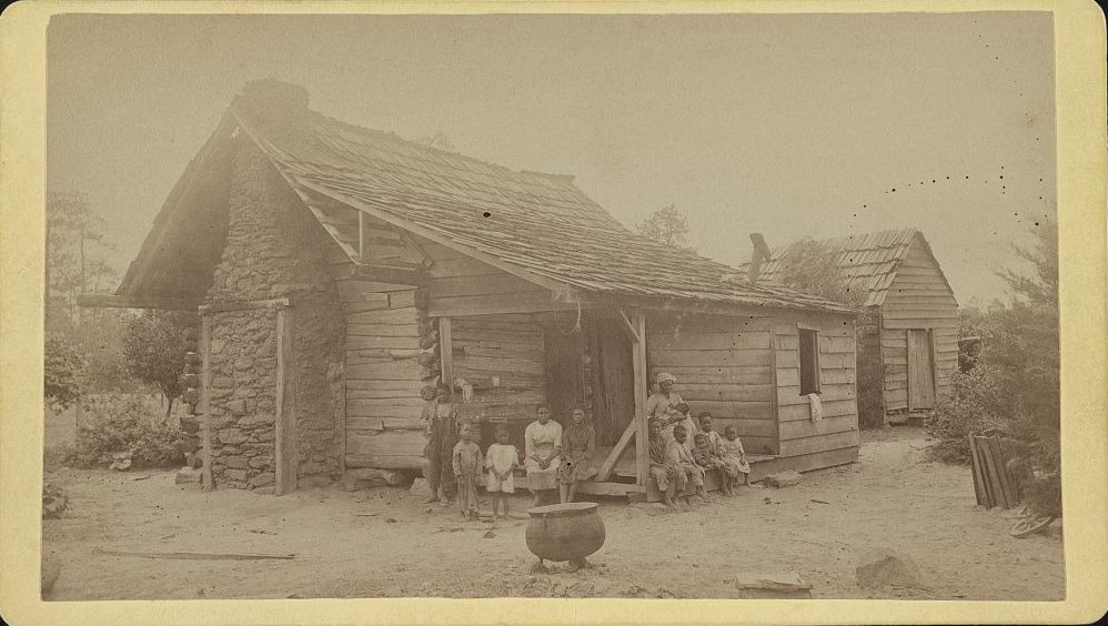 A Black family posed outside their hewn log home in the nineteenth century.