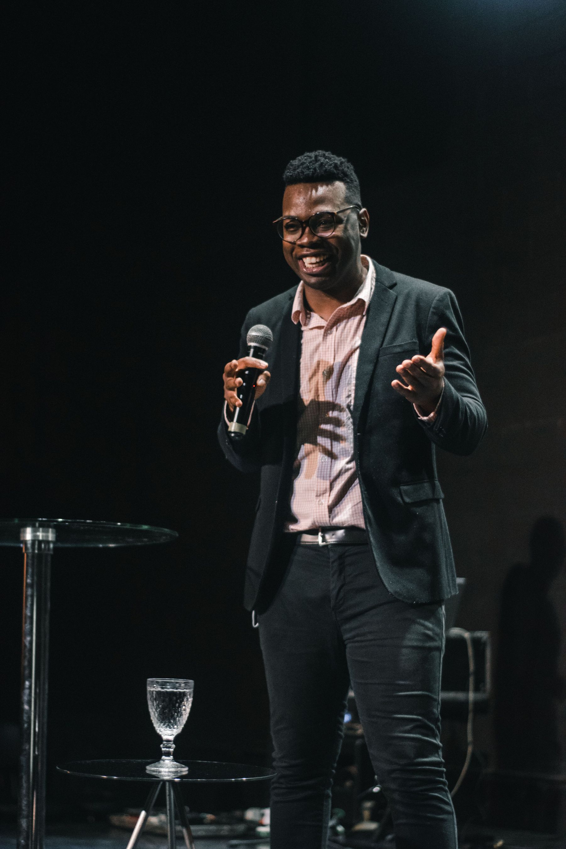 A speaker in a suit addressing an audience from a stage with a handheld microphone.
